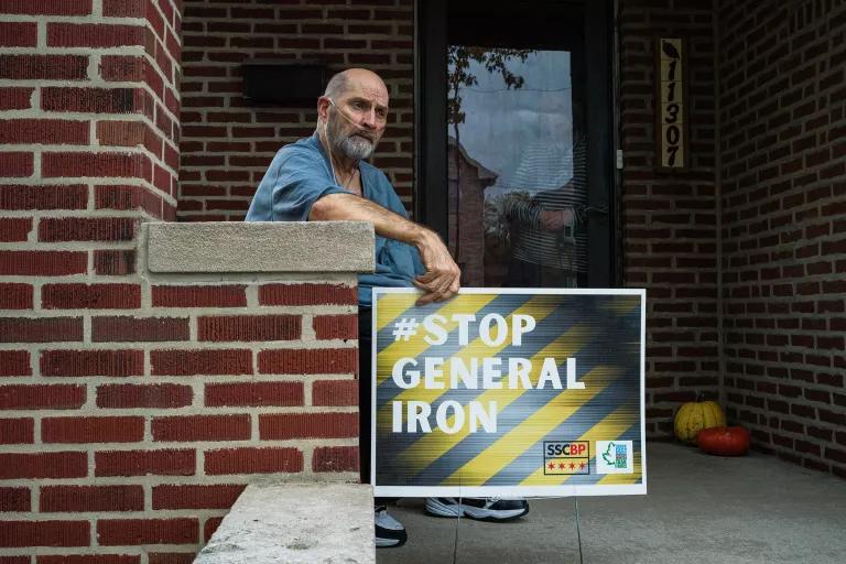 A man sits on a the stoop of a brick building with a poster that reads "#Stop General Iron"