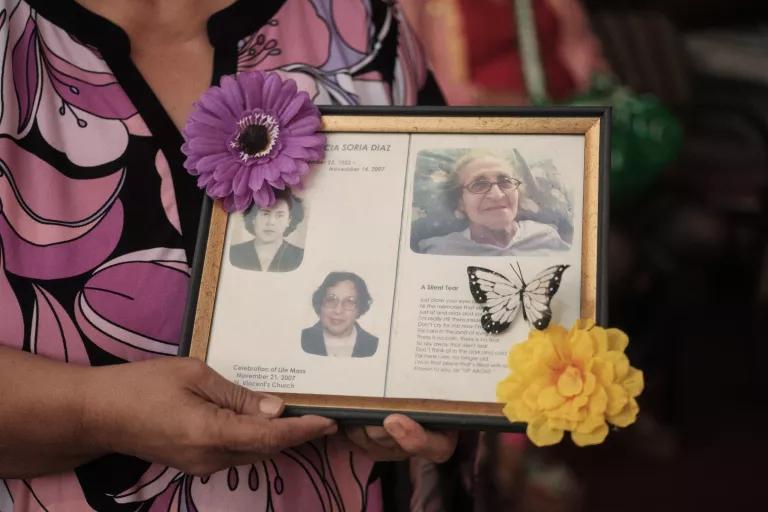 A woman holds a framed plaque that has three photos of a woman and is decorated with artifical flowers and a butterfly
