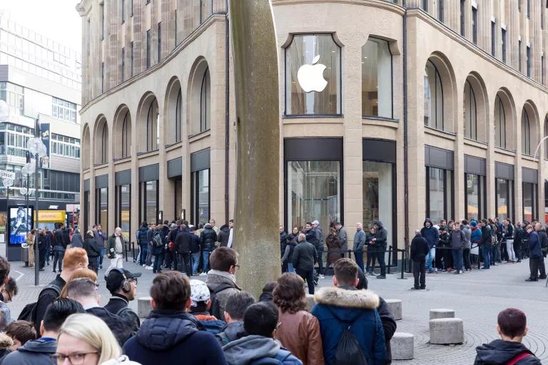 A long line of people standing on a city sidewalk outside of an Apple store