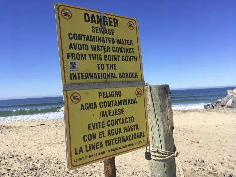 A yellow sign stands in the sand on a beach reads in both English and Spanish, "Danger: Sewage Contaminated Water; Avoid water contact from this point south to the international border"