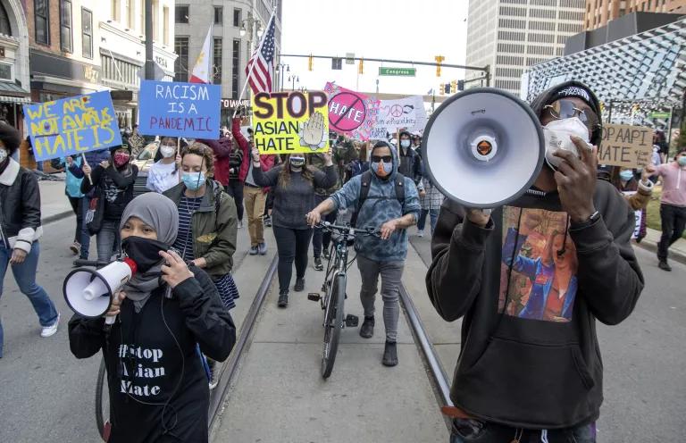 A protest with protestors in the front holding megaphones, and others in the back holding signs that say "Stop Asian Hate" and "Racism Is a Pandemic"
