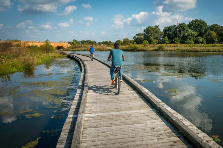 Cyclists ride on a boardwalk over a calm body of water