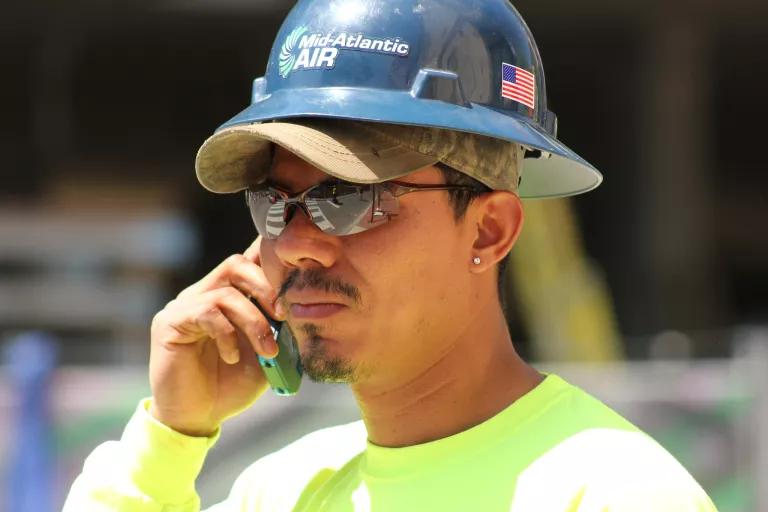 Close up of a construction worker talking on a cell phone. He is wearing wrap around sunglasses and a hard hat over his ballcap. 