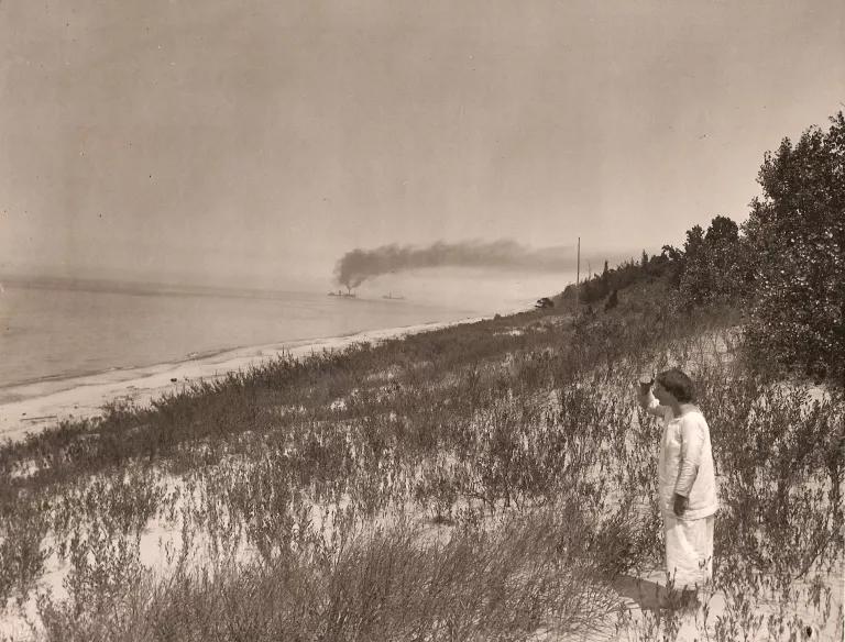 A sepia-toned photo shows a woman standing in tall grass on a dune with smoke from a ship in the ocean rising in the distance