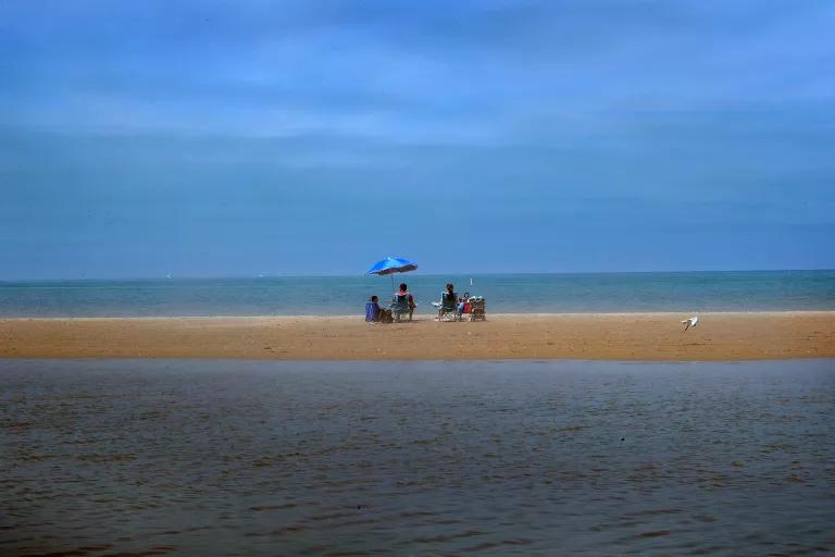 People sit on beach chairs with an umbrella on a narrow strip of sand in calm waters