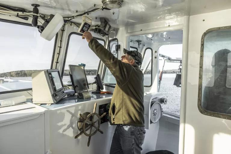 A man stands in the wheelhouse of a boat and reaches up toward a small digital display