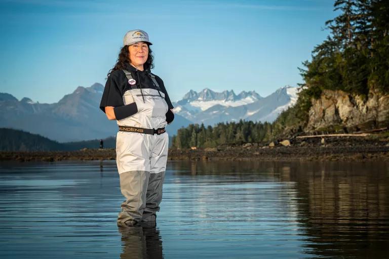 A woman in waders stands in shallow waters with mountains in the background