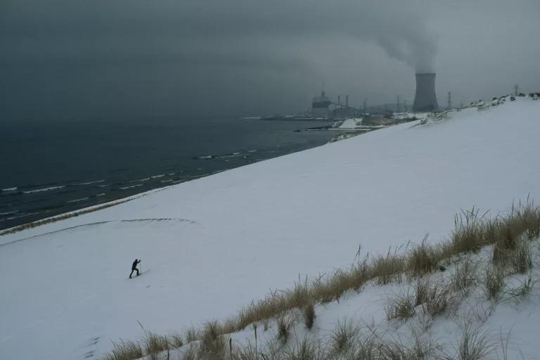 A lone skier moves across a snow-covered dune under cloudy skies with a power plant cooling tower in the distance