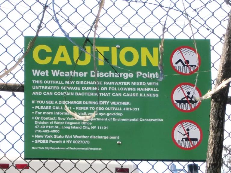 A sign that hangs on a chain link fence is titled "Caution: Wet Weather Discharge Point"