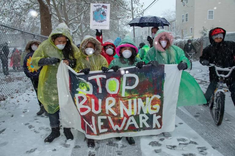 A group of protesters walk on a snowy street and hold a sign that reads "Stop Burning Newark"