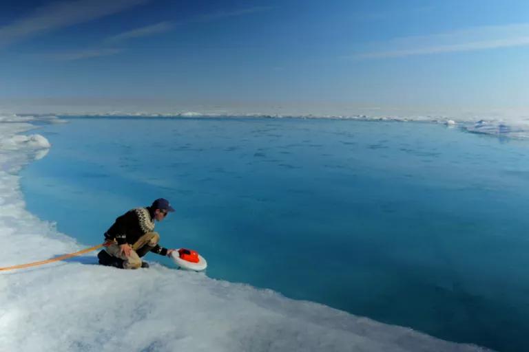 A man sits on a swath of ice at the water's edge, holding a red and white disk above the water