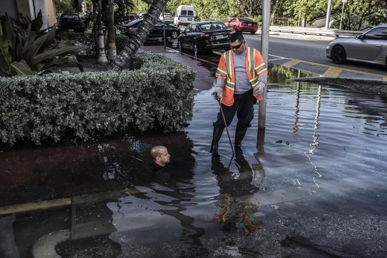 A worker stands in ankle-deep floodwater on a city street, while another worker's head is visible just above the water as he reaches down into it