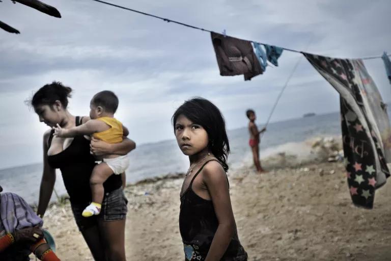 A child stands on a beach next to a clothesline while a woman holding an infant stands nearby