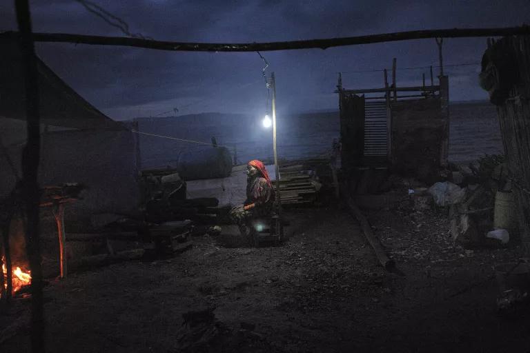 A woman sits beneath a single light bulb hanging from a pole in a clearing surrounded by tents and small structures