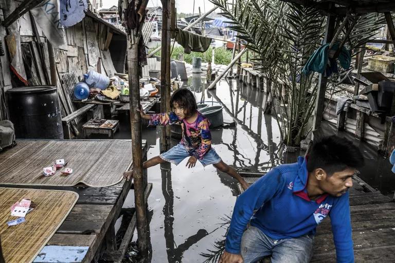 A young girl stands with one foot on a platform in front of a wooden house and the other foot on a lower platform, with floodwaters below her