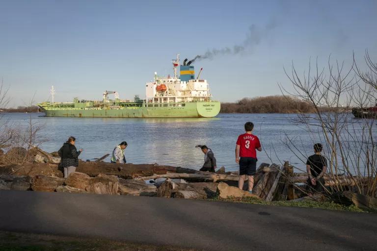 People stand on a riverbank as a cargo ship passes by in the river