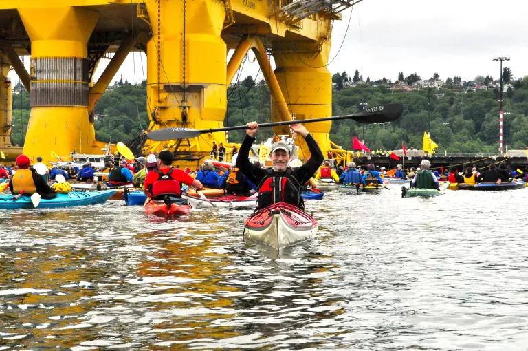A person waring a cap on a kayak in the water holding up the paddle, with other kayakers on the water and a yellow oil rig in the background