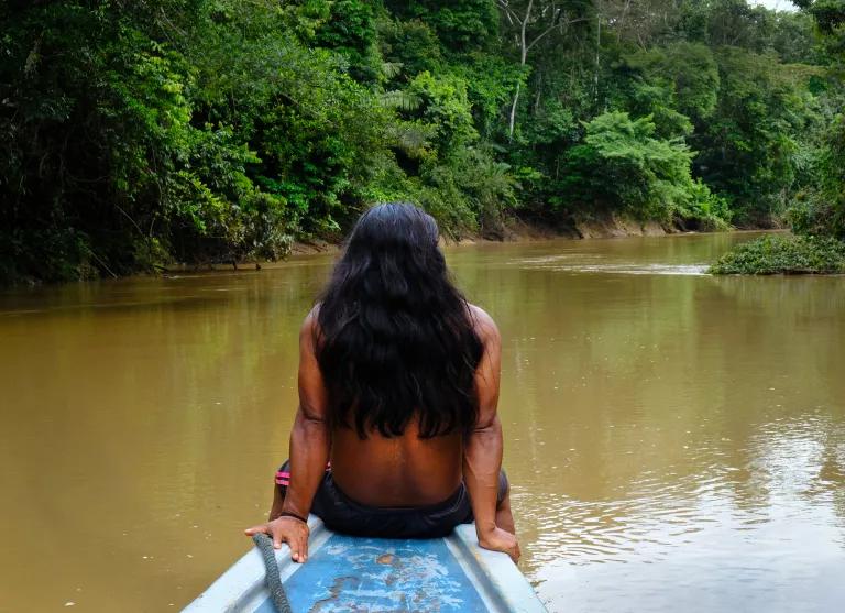 A person sits on the end of a longboat on a brown river