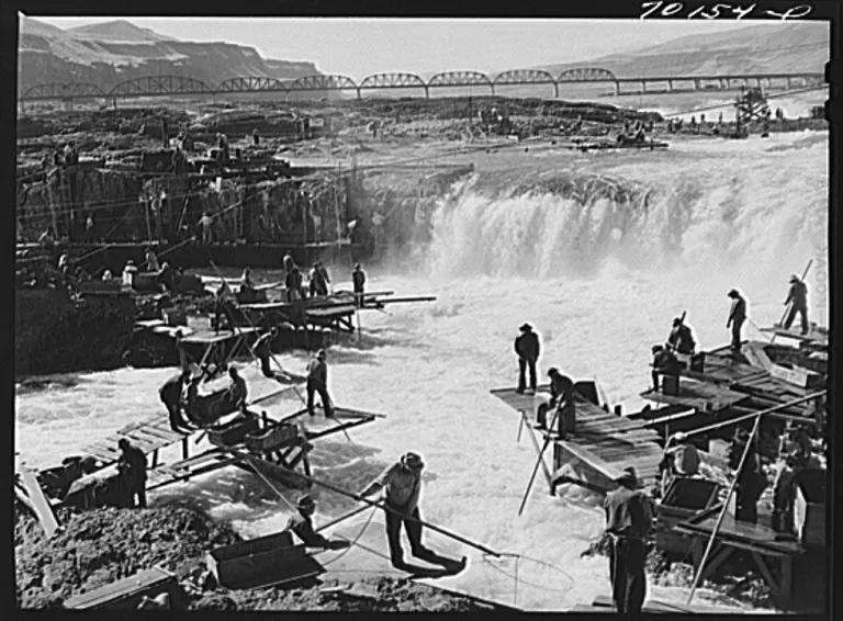 A black-and-white shows men standing on platforms fishing in a turbulent river with a waterfall in the distance