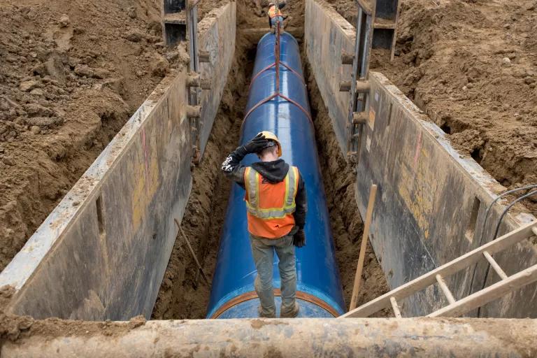 A worker stands above a large pipe that sits inside a deep trench