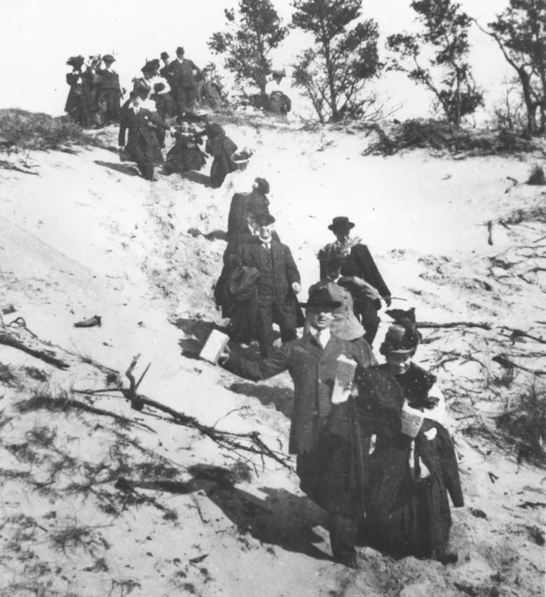 A black and white photo of men and women in dress clothing walking down a large dune