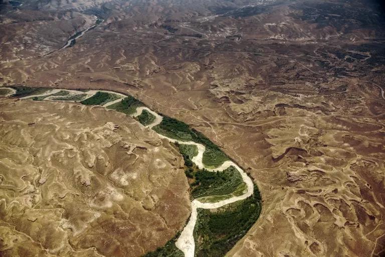 An aerial view of a stretch of a river snaking through a narrow green valley with brown mountains on both sides