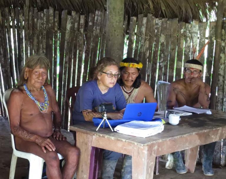 A woman sits at a table in a hut and works on a laptop while three men sit near her