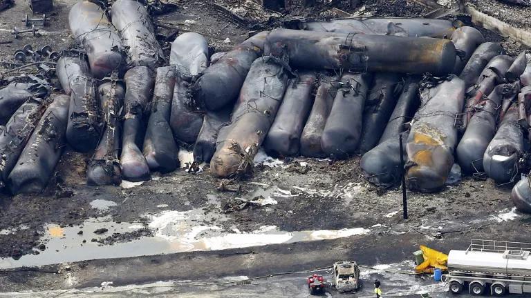 More than 20 burned and charred oil tanker train cars lie next to and on top of each other with emergency vehicles nearby