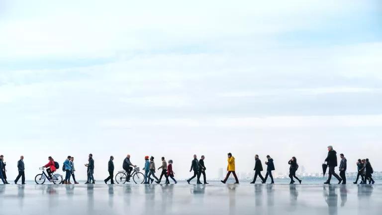People walk on a paved walkway near a body of water with a city skyline in the background