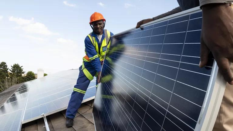 A man in an orange hard hat holds a solar panel on a roof