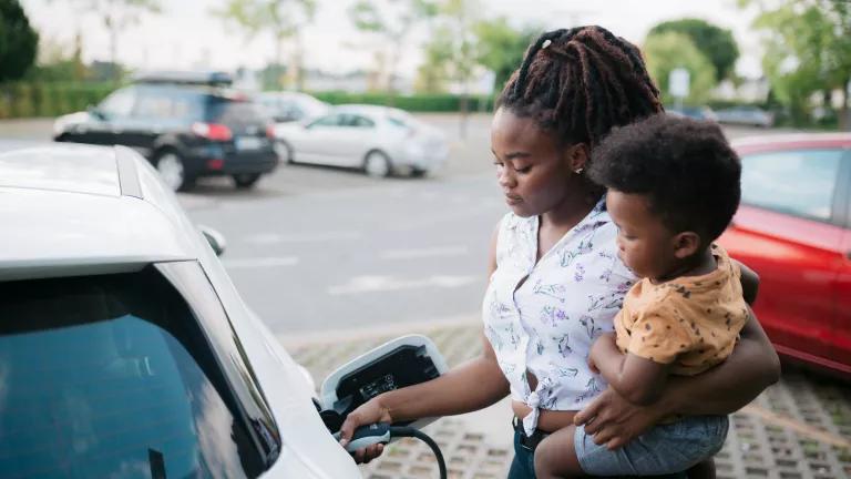 A mother holding her baby son as she charges her electric car at a charging station.