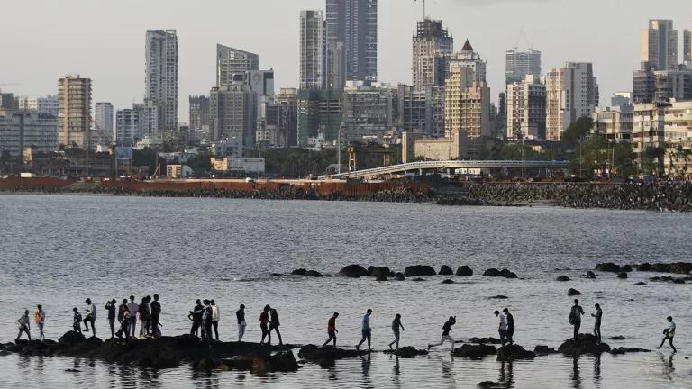 People walking on rocks in a bay, with the Mumbai cityscape visible in the background.