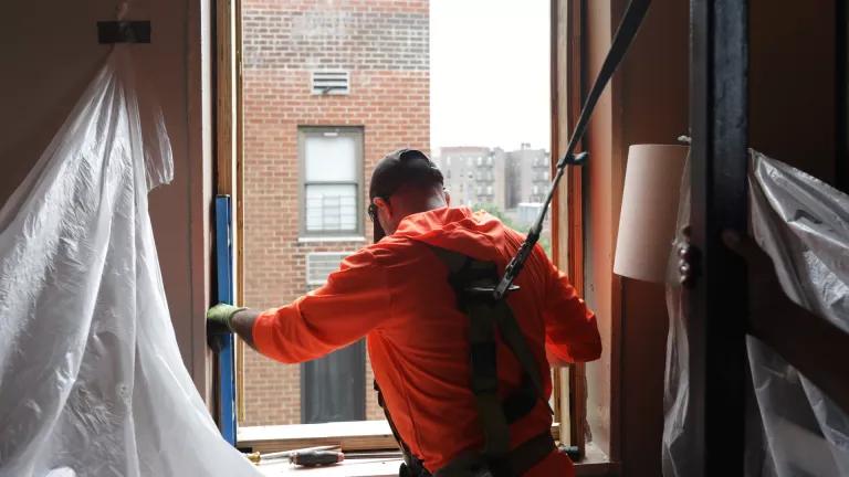 A worker in an orange jacket looking out an open window, getting ready to install a new energy efficient window in an apartment