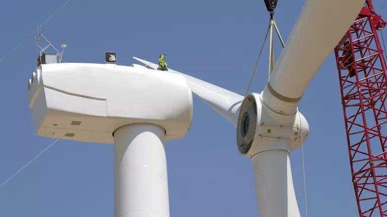 A worker stands on top of a wind turbine and guides a crane attaching the rotor and blades.