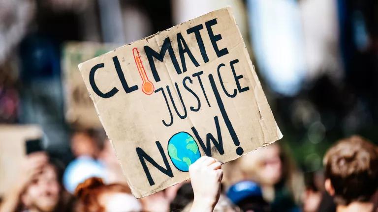 A hand holding up a sign on brown cardboard that reads "Climate Justice Now!"