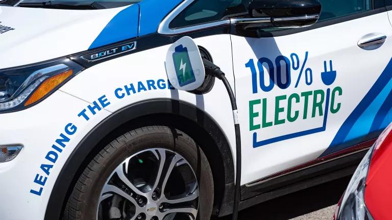 A vehicle painted with the words "100% Electric" is plugged into a charger
