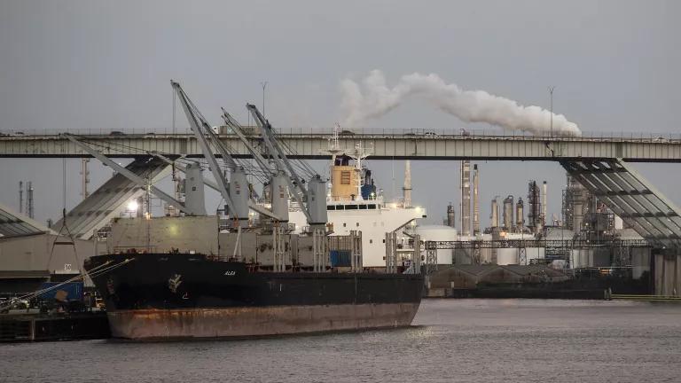 A large gray tanker ship with a bridge and refinery stacks in the background