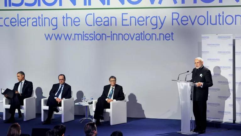 A man stands at a lectern next to three other men sitting in chairs on a stage 