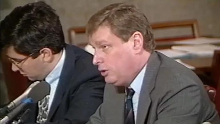 A man in a suit sitting and speaking into a microphone, with another man in a suit and glasses next to him
