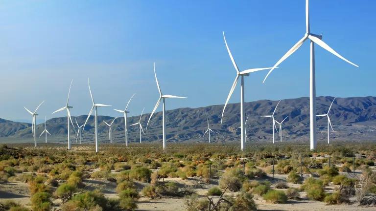A row of wind turbines stand on a desert plain with mountains in the backgound