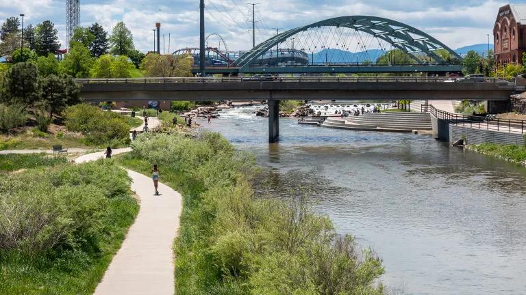 A bridge spans a river beside a park in Denver, Colorado. People are running on a riverside track.