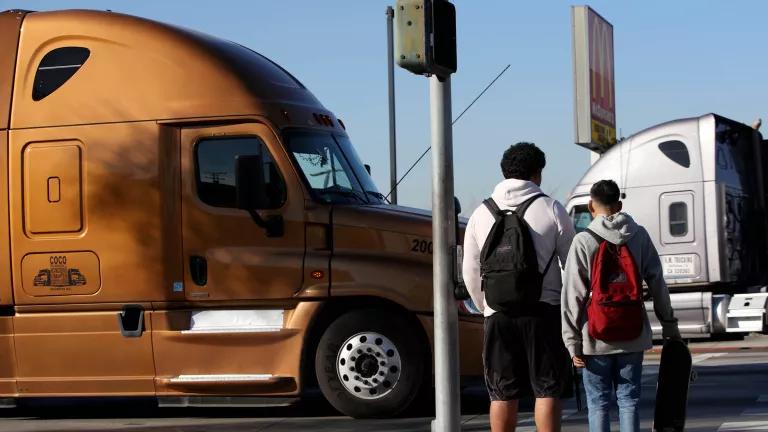 A view from behind of two teenage students waiting to cross an intersection with heavy semi-trailer truck traffic