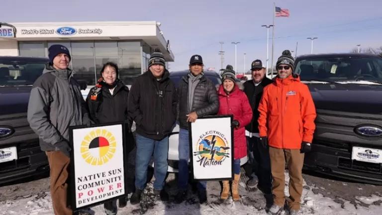 Seven people in winter coats standing in front of parked cars facing the camera, one person with a red jacket holding a sign that says Electric Nation