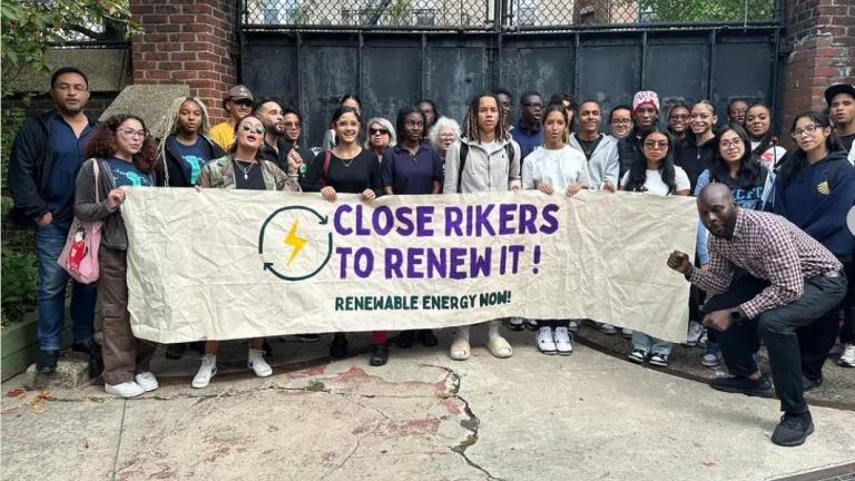 A group of people standing with a large banner that says "Close Rikers to Renew It! Renewable Energy Now"