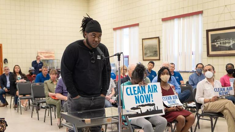 Qiyam Ansari speaks into a microphone at a public hearing in front of a seated room of people. Attendees in the front row hold signs that read 'Clean Air Now!'