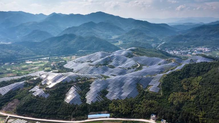 Solar panel arrays cover a hilltop, surrounded by trees and taller mountains