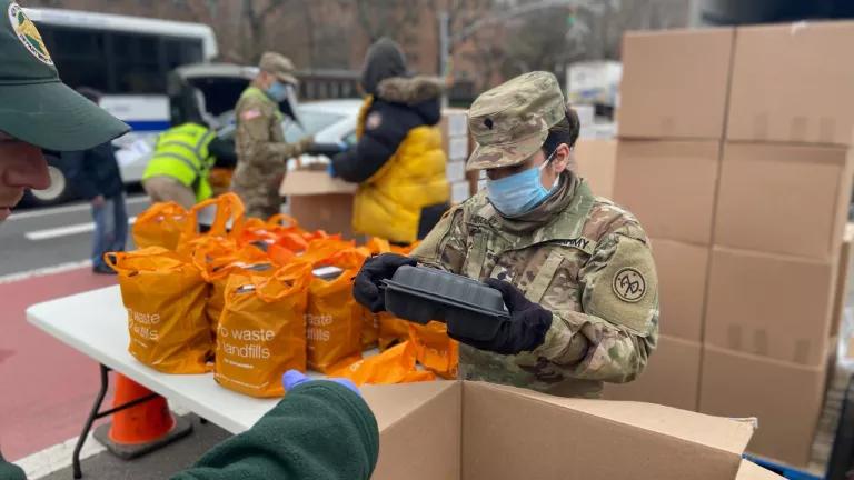 A woman wearing Army fatigues, gloves, and a mask stands at an outdoor table packing pre-made meals into reusable bags.