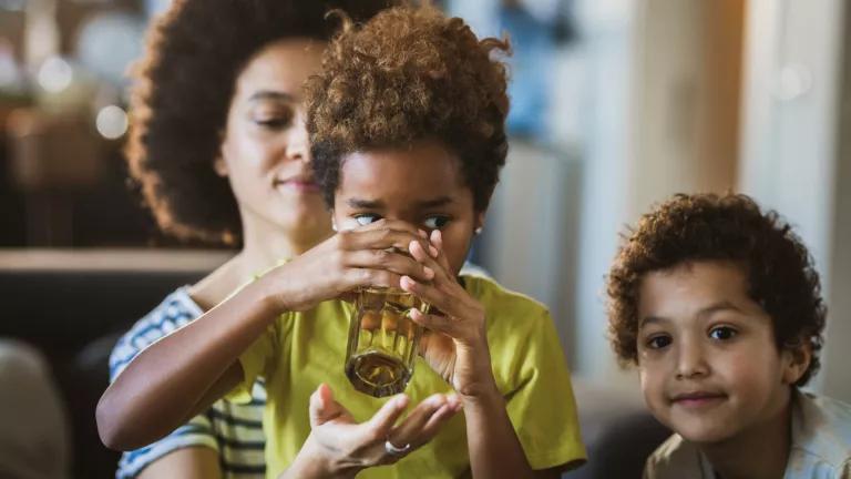 A child drinking water out of a glass an and adult behind them holding up a hand below the glass