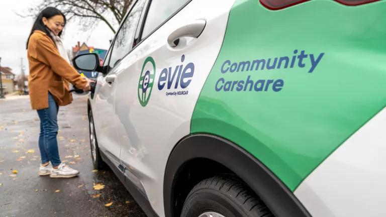 Asian American woman with jeans and a tan coat plugs a charger into an Evie Carshare vehicle (white Chevy Bolt with an Evie Carshare decal).