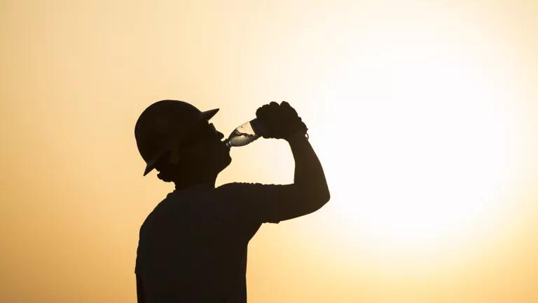 A person with a round hat drinking a bottle of water with the sun in the background